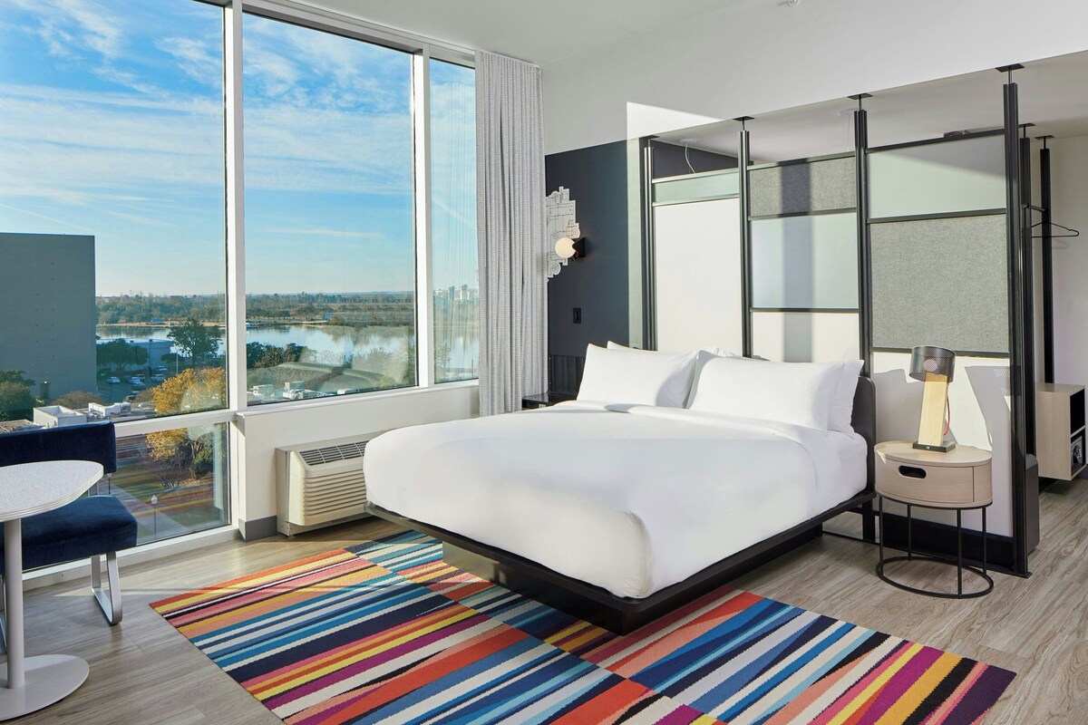 A modern hotel room with a large bed, a view of a cityscape, and a colorful striped rug.
