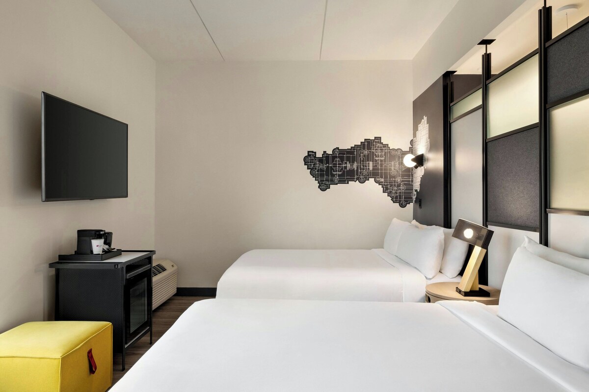 A hotel room with two beds, a flat-screen TV, a coffee machine, a small table, and a decorative wall piece.
