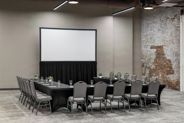 Un salón de conferencias con una larga mesa, sillas, una pantalla de proyector y una pared de ladrillo.
