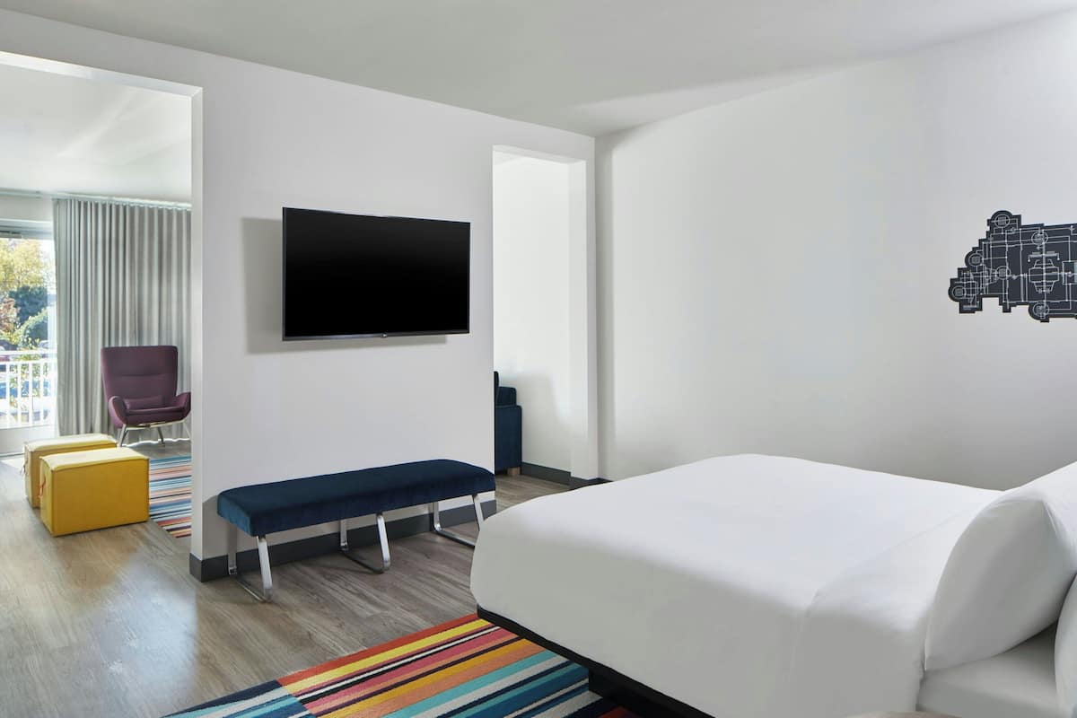 A modern hotel room with a large bed, a flat-screen TV mounted on the wall, a bench, a colorful striped rug, and a view of the outdoors through a large window.