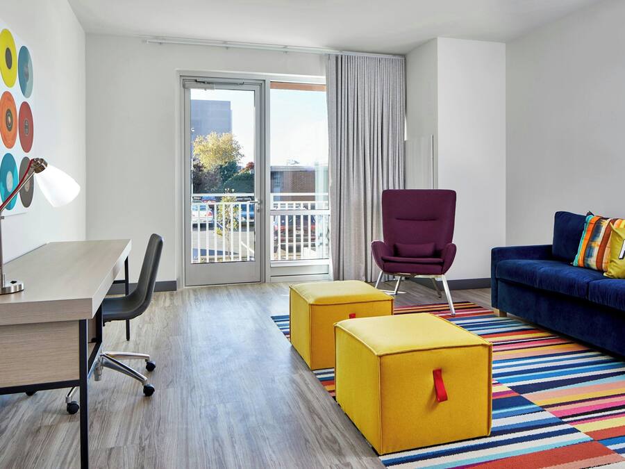 A modern hotel room with a desk, two yellow ottomans, a purple armchair, a blue sofa with a colorful pillow, and a large window with a view of the outside.