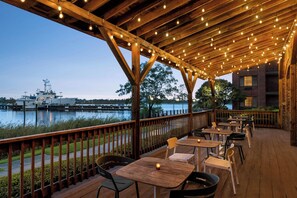 An outdoor deck with wooden tables and chairs, overlooking a body of water and a docked ship. - Aloft by Marriott Wilmington at Coastline Center (Wilmington)