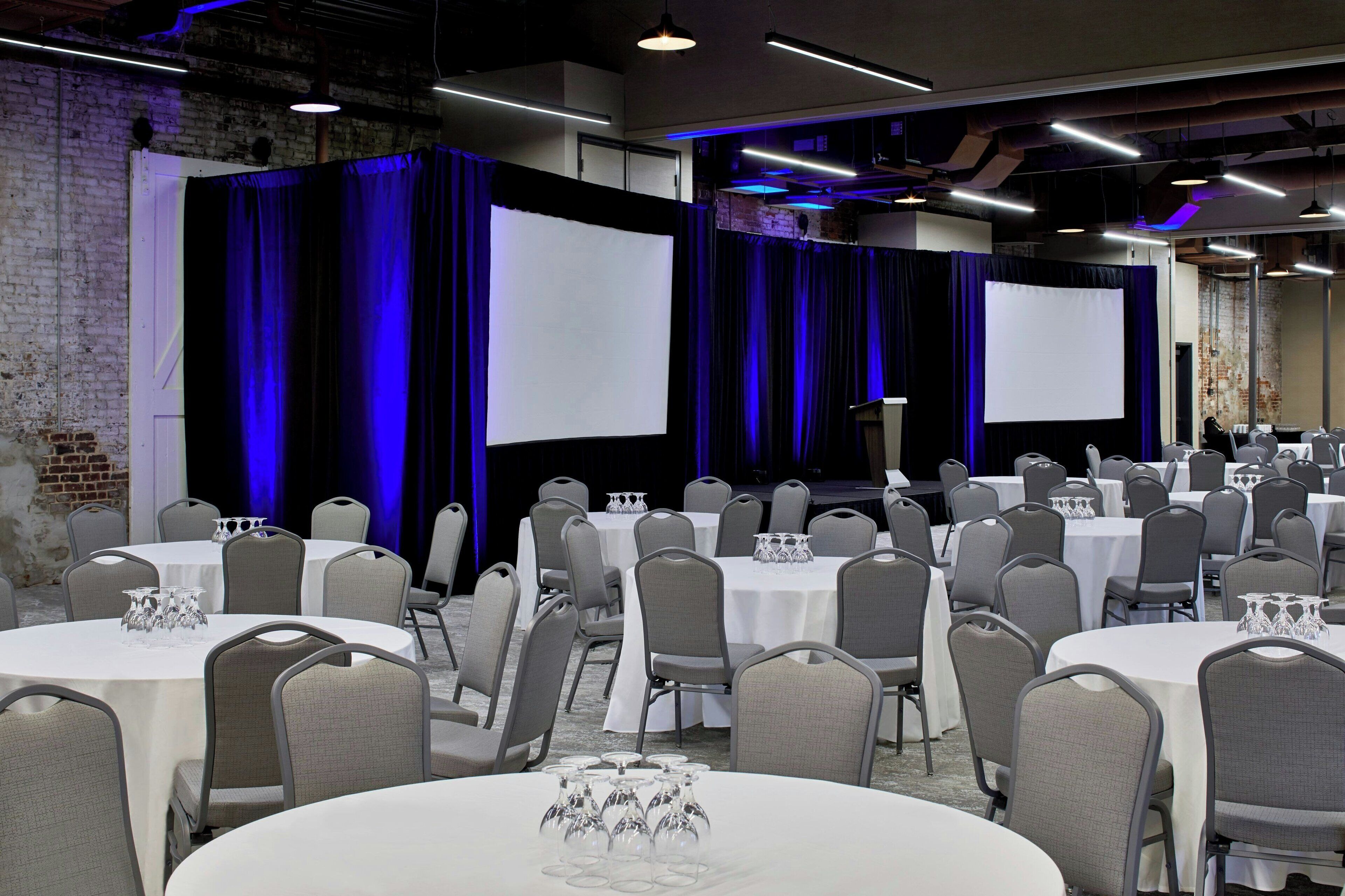 Une salle de conférence équipée de plusieurs tables et chaises, de grands écrans et de rideaux bleus.