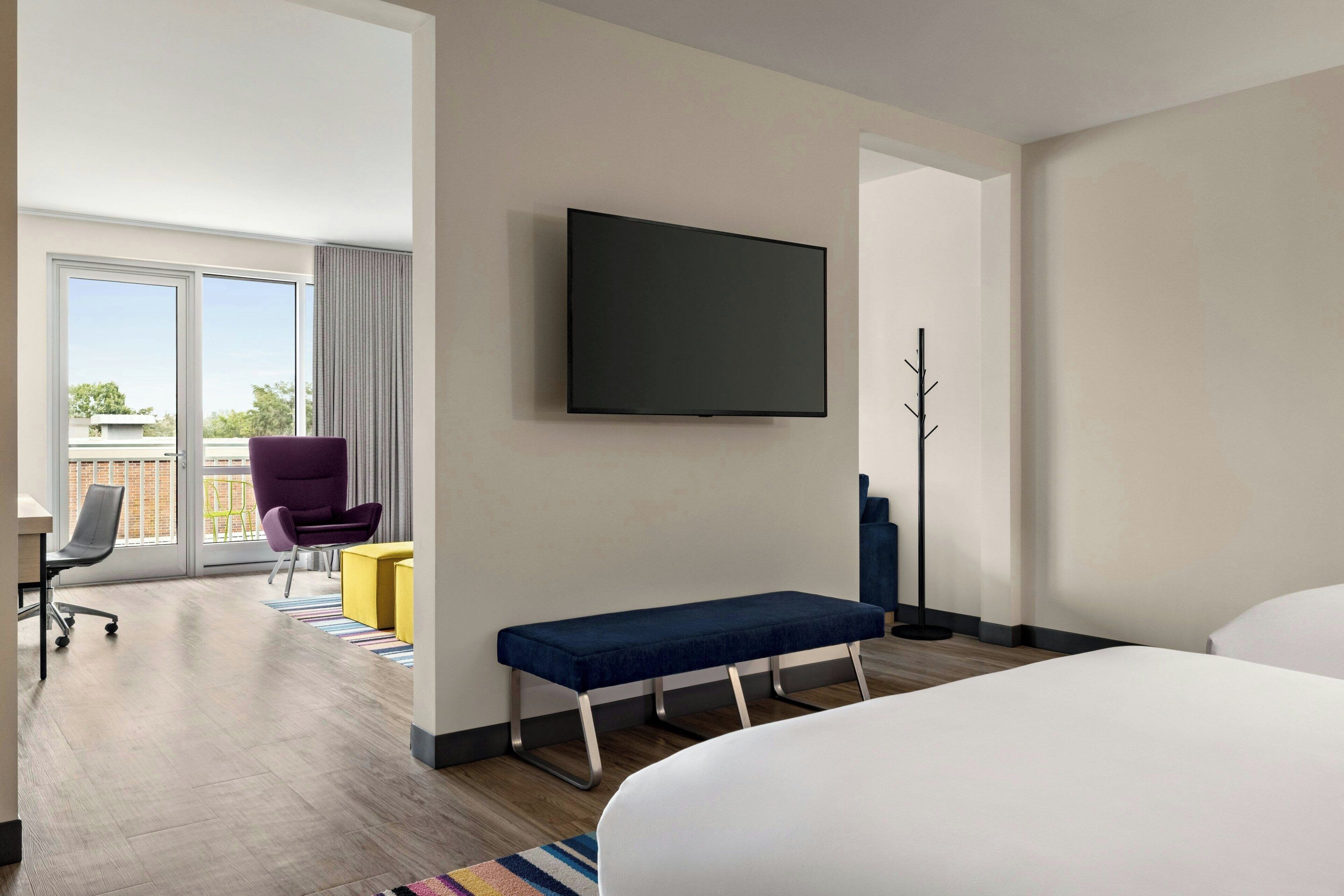 A modern hotel room with a large bed, a flat-screen TV mounted on the wall, a bench, a chair, and a desk.