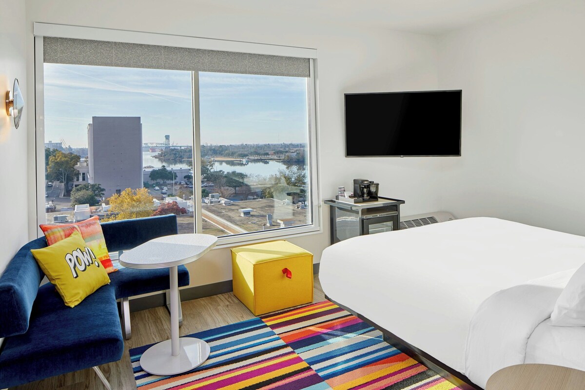 A hotel room with a large window offering a view of a cityscape and water, a bed, a small table, a sofa with colorful pillows, and a flat-screen TV mounted on the wall.