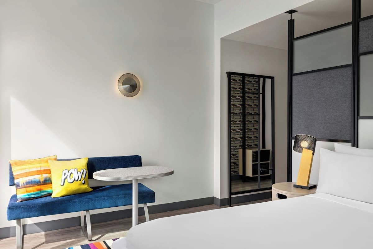 A modern hotel room with a bed, a round table, a blue sofa with colorful pillows, a wall-mounted light, and a sliding door closet.