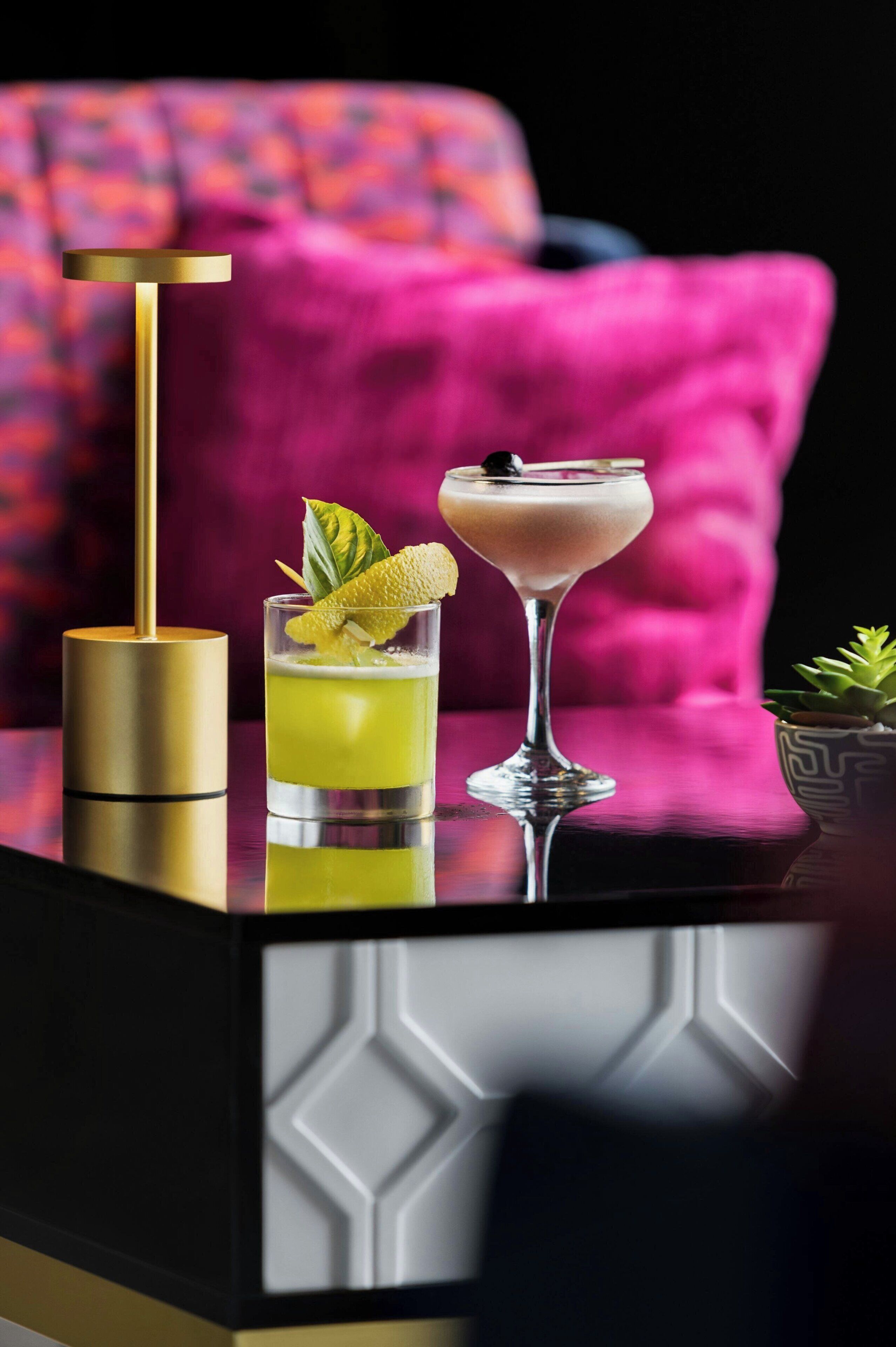 Deux cocktails sont posés sur une table noire réfléchissante, dont le piédestal est doré. On aperçoit également un objet doré et, à l’arrière-plan, une chaise rose recouverte de tissu.