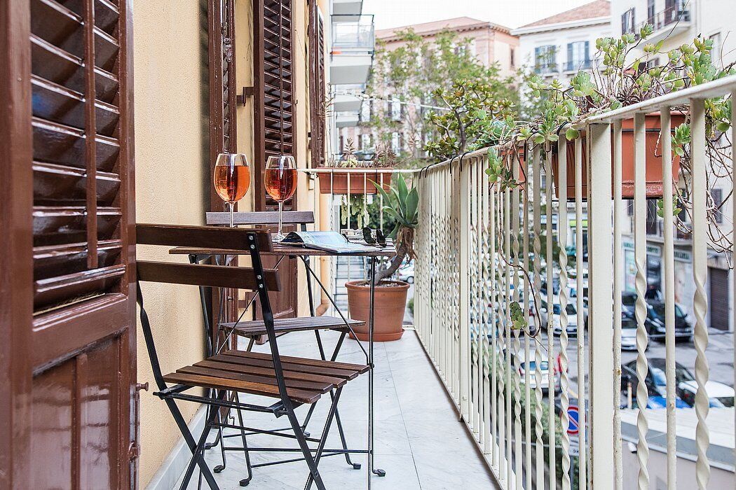 A balcony with a wooden table and chairs, two glasses of red wine, and potted plants.