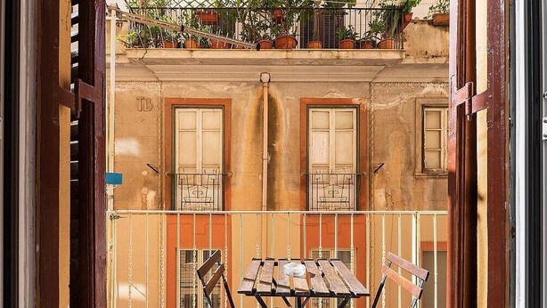 A balcony with a wooden table and chairs, potted plants, and a building facade with windows and a door.