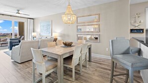 A spacious living area with a wooden dining table, comfortable seating, a chandelier, and a view through large windows. - Ariel Dunes II 509 - Gulf View, Heated Community Pool, Steps to the Beach! (Miramar Beach)