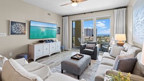 A spacious living room with a flat-screen TV, a ceiling fan, and a view of the city through large windows. - Ariel Dunes II 509 - Gulf View, Heated Community Pool, Steps to the Beach! (Miramar Beach)