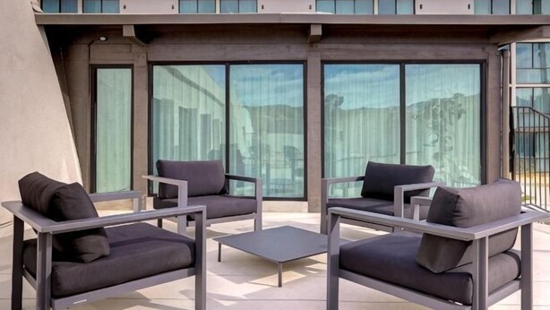 A modern outdoor seating area with sofas and a coffee table, large windows, and a building facade in the background. - DoubleTree by Hilton Palmdale (PALMDALE)