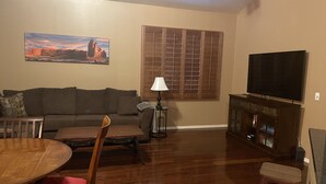 A living room with a wooden floor, a sofa, a TV on a cabinet, a painting on the wall, and a window with blinds. - Beautiful centrally located townhome in The Gardens, Gilbert (Gilbert)