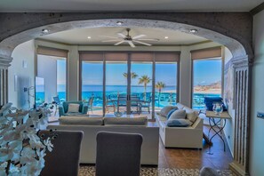 A spacious living area with a large window overlooking a pool and beach, a ceiling fan, and a modern interior design.