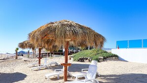 A beach with white lounge chairs, a thatched umbrella, and a white building in the background. - Esmeralda Beach Resort 2 Bedrooms Condo B 502 (Puerto Peñasco)