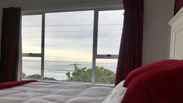 A bed with a white textured cover and red pillows, a window with red curtains, and a view of the ocean. - Soothing Sounds (Otago)