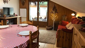 A cozy, well-lit attic room with a dining table set for two, a comfortable couch, and a small TV. - 3-room cabin apartment, south-facing balcony, 5 pers., private parking (Les Contamines-Montjoie)