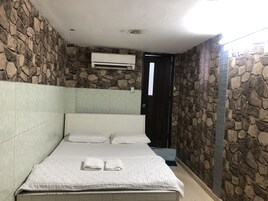 Quarto com cama, almofadas brancas, ar condicionado fixado na parede e uma porta.