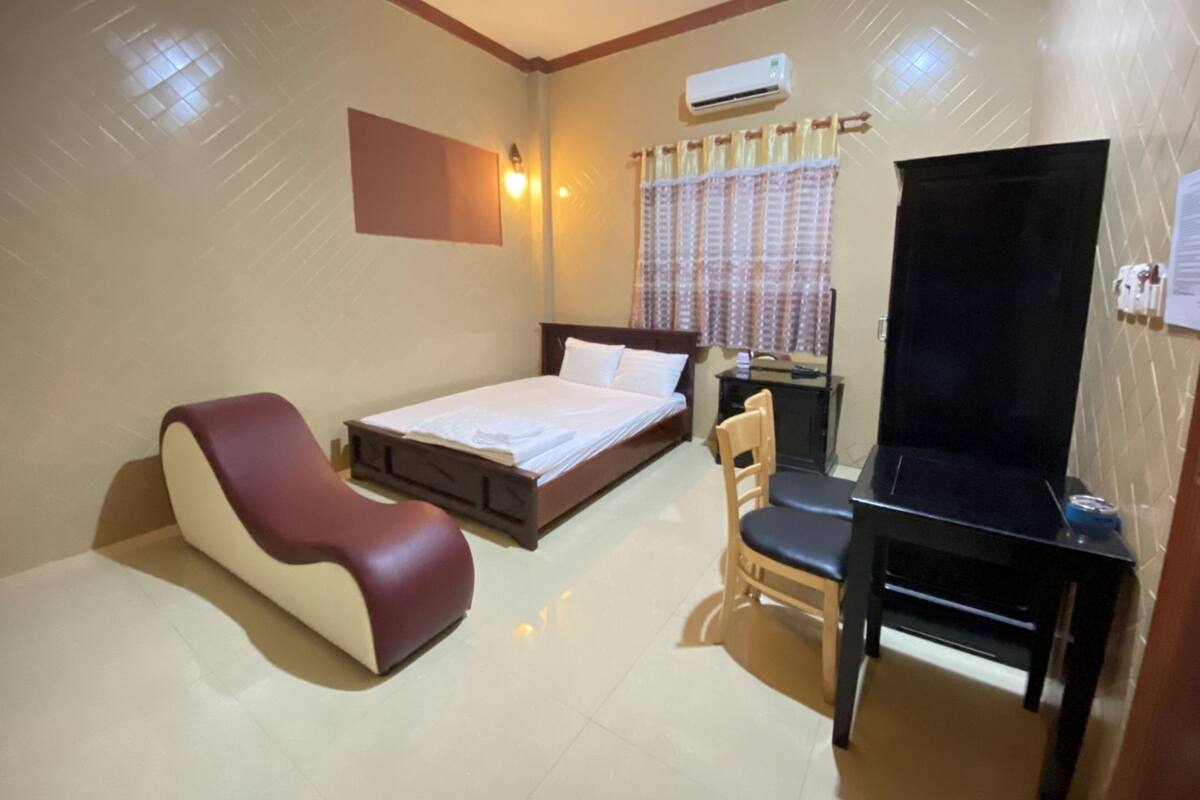 A hotel room with a bed, a desk with a chair, a TV, and an air conditioning unit.