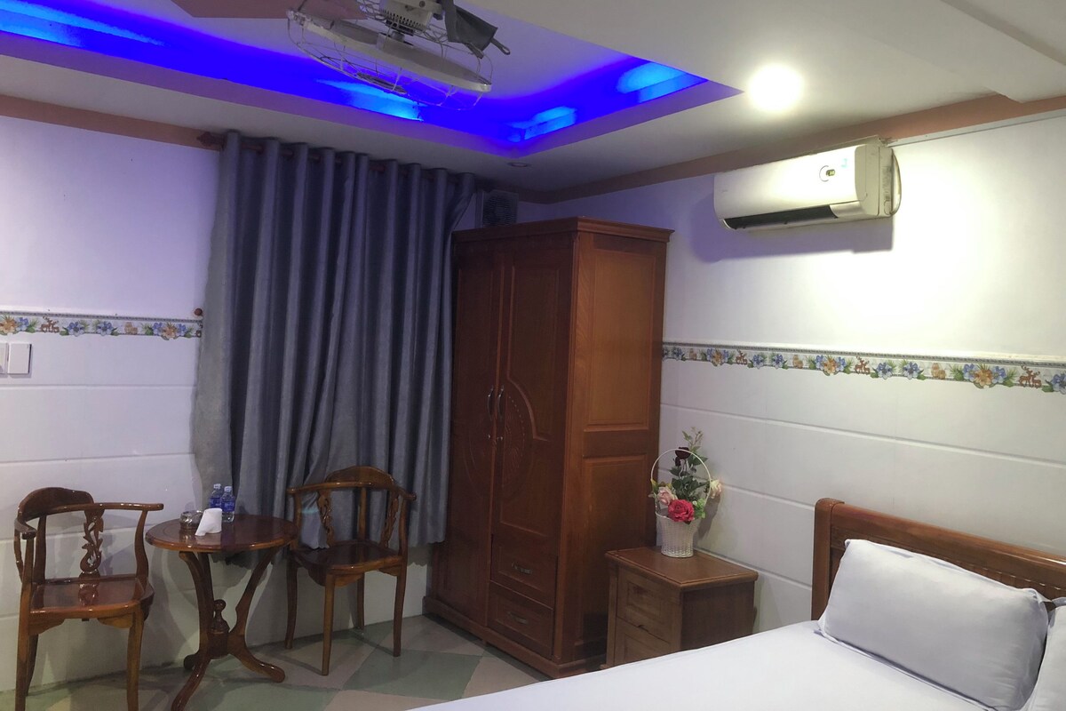 Een hotelkamer met een bed, een houten garderobe, een bureau met stoelen, een kleine tafel met een bloevas en een plafondventilator.