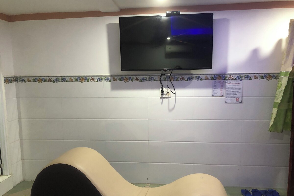 Una habitación con un televisor montado, un enchufe en la pared, una pared con azulejos y un borde decorativo, y un par de pantuflas azules en el suelo.