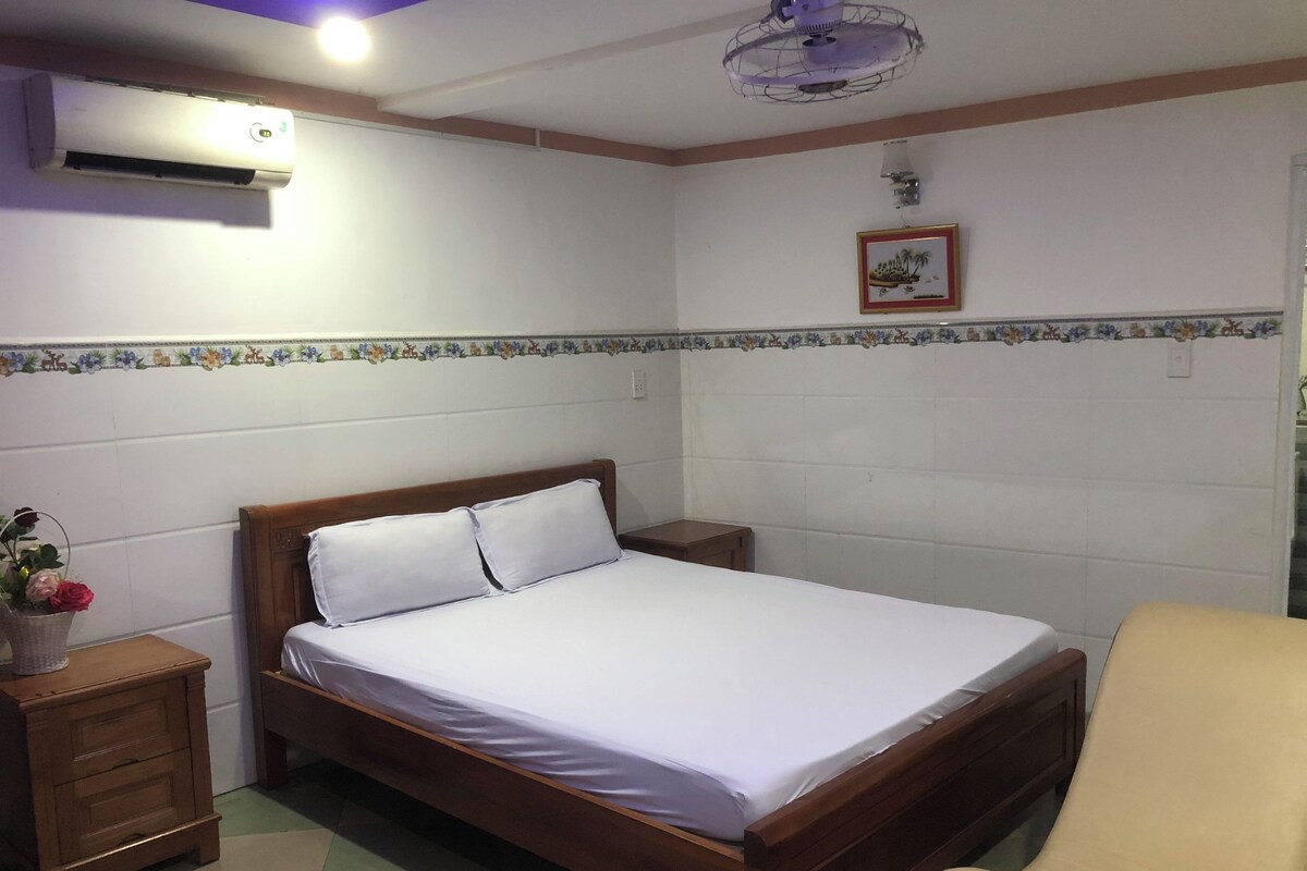 Un dormitorio con una cama de madera, una mesita de noche, una silla, un aire acondicionado instalado en la pared y un ventilador de techo.