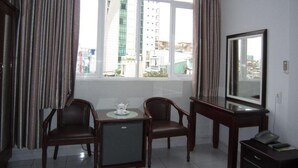 Habitación de hotel con un ventanal grande, dos sillas, una mesita con tetera, un espejo y un teléfono sobre una mesita auxiliar.