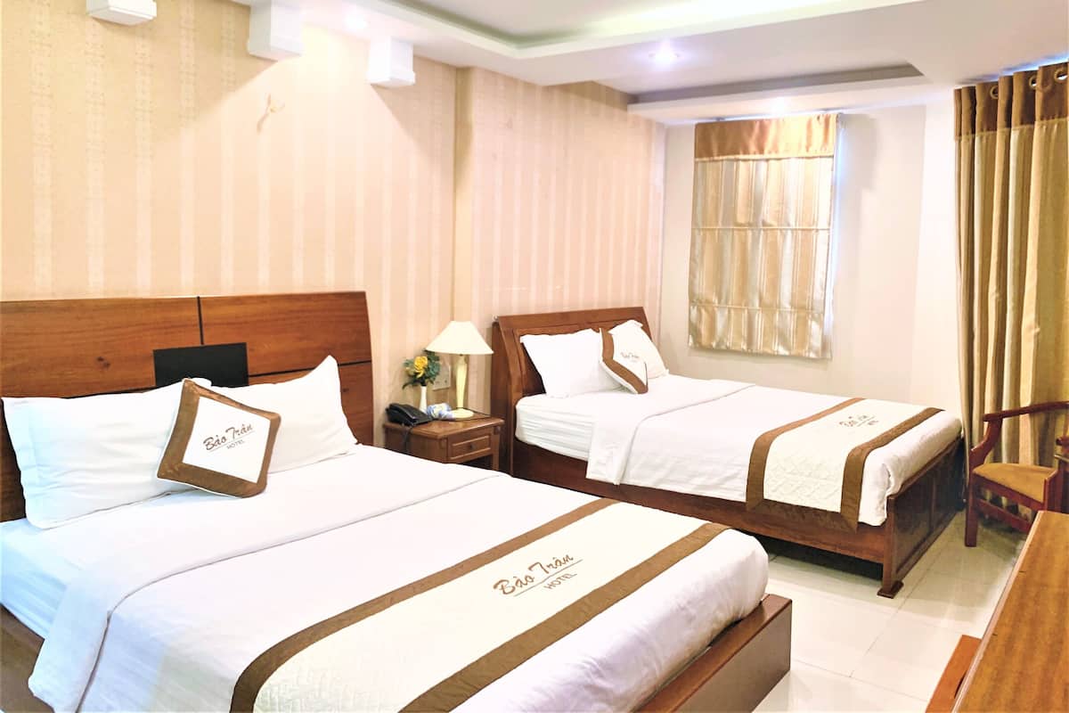 Ein Hotelzimmer mit zwei Betten, beide mit Holz Kopfteilen, weißer Bettwäsche und braunen Zierelementen. An einem Bett befindet sich ein Nachttisch mit einer Lampe und einem Telefon, neben dem anderen Bett steht ein Sessel.