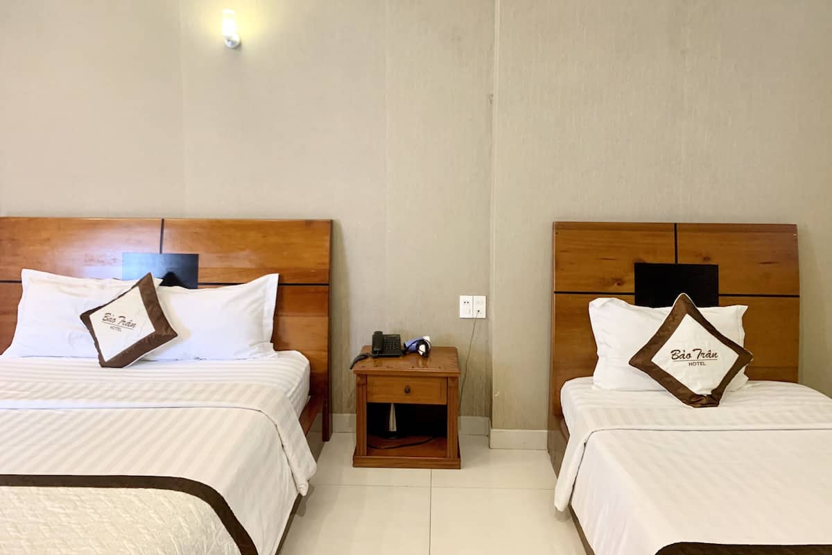 Ein Hotelzimmer mit zwei Betten, jedes mit einem hölzernen Kopfende und einem Kissen mit dem Namen des Hotels. Dazwischen befindet sich ein Nachttisch mit einer Lampe und einem Telefon.