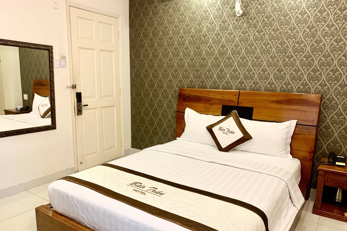 Ein Hotelzimmer mit einem großen Bett, einem Holzkopfstück, einem Nachttischchen mit Telefon und einem Spiegel an der Wand.