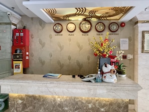 A hotel reception desk with clocks displaying times from various cities, a Coca-Cola vending machine, and a decorative vase with flowers.