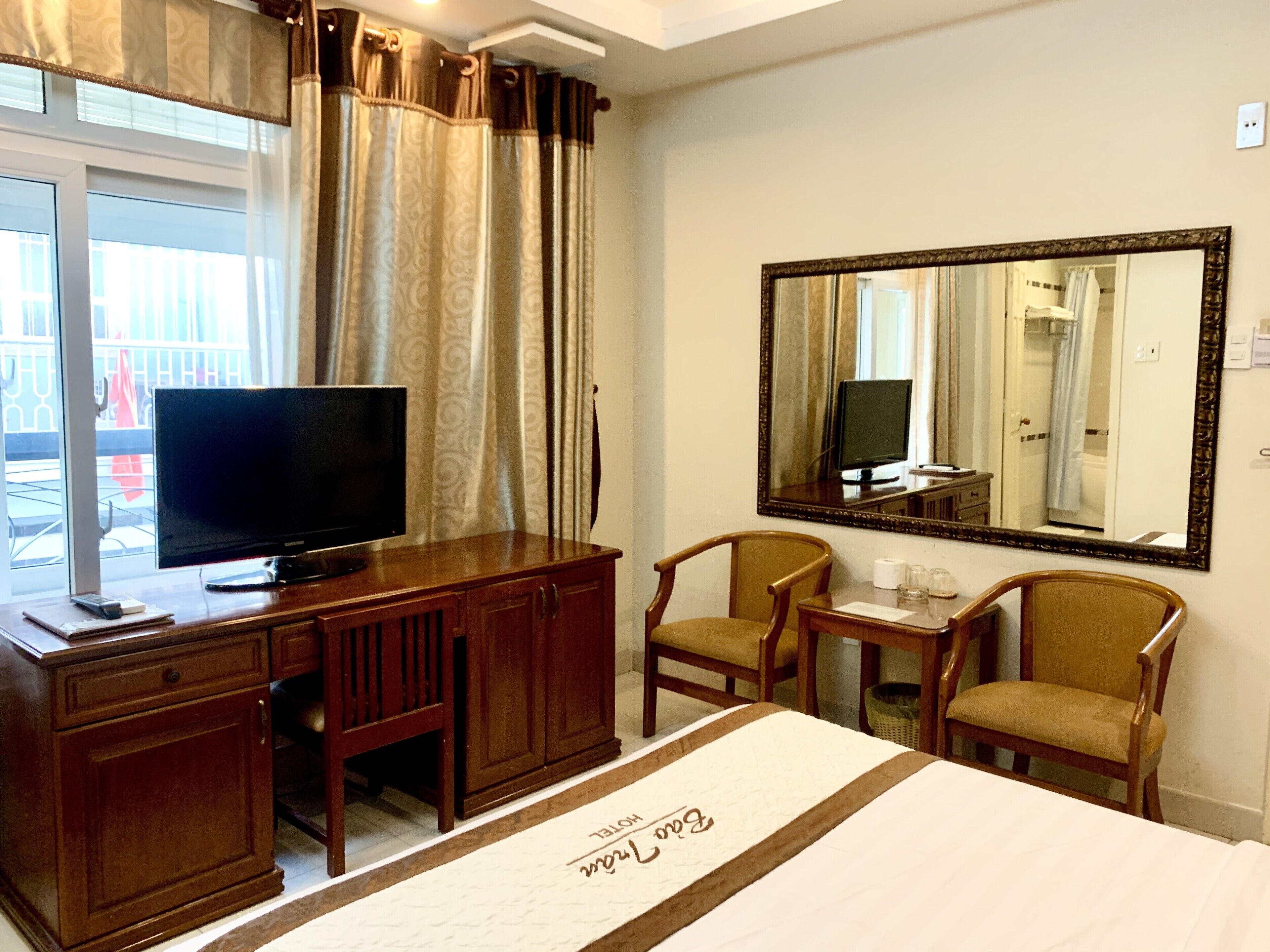 A hotel room with a TV on a wooden desk, two chairs, a mirror, and a bed with a decorative bedspread.