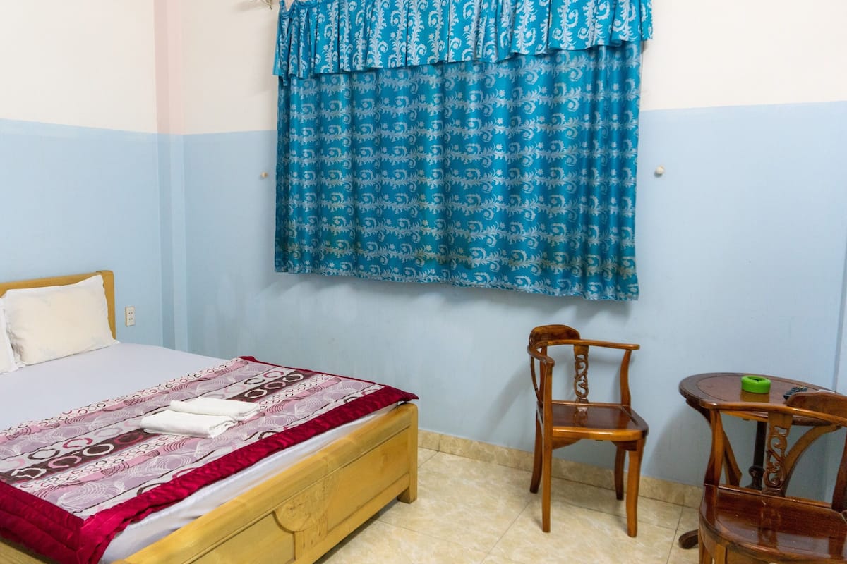 Ein Schlafzimmer mit einem hölzernen Bett, einem Sessel, einem kleinen Tisch und einem Fenster mit gemusterten Vorhängen.