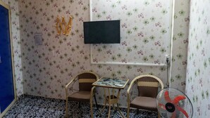 A small room with floral wallpaper, a television mounted on the wall, two wicker chairs, a small table, and a standing fan. - Khanh Vy Hotel Binh Tan (Ho Chi Minh City)