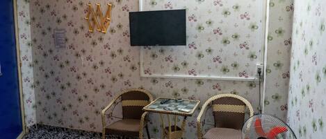 A small room with floral wallpaper, a television mounted on the wall, two wicker chairs, a small table, and a standing fan.