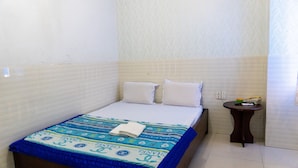 Een klein, netjes opgemaakt bed met een blauw-witte bedspreide en twee kussens, een klein nachtkastje met een telefoon en een kopje, en een wand met een subtiel bloemenpatroon.