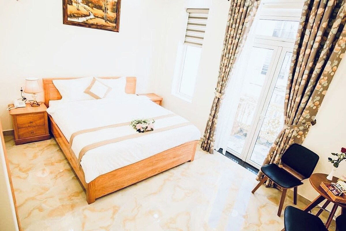 A neatly arranged bedroom with a large bed, bedside tables, a chair, and a small table with a vase.