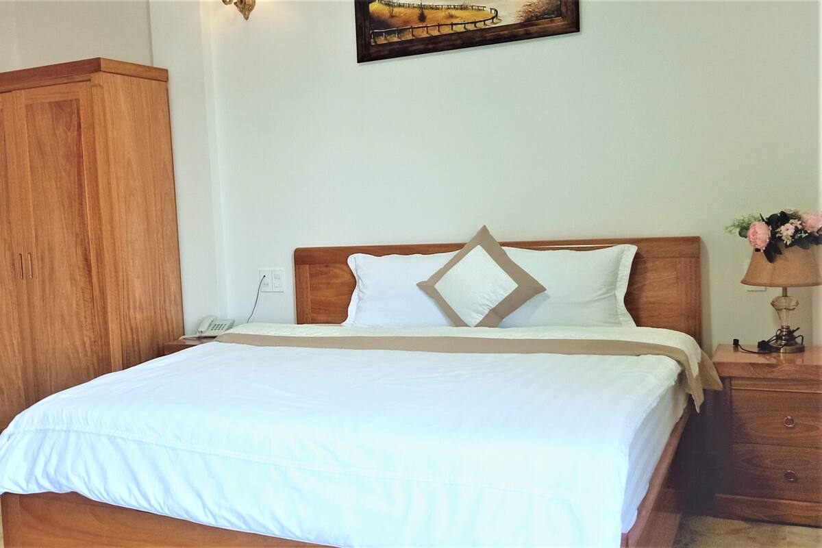 A neatly made bed with white bedding and a wooden headboard. There is a wooden nightstand with a lamp and a vase of flowers to the right. A wooden wardrobe is visible to the left of the bed.