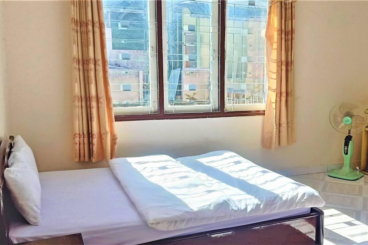 A single bed with white bedding in a room with a window, a standing fan, and a small table.