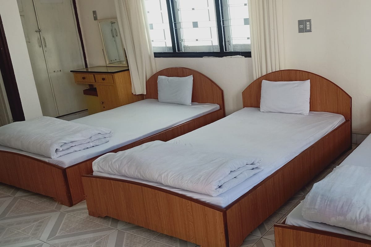 A hotel room with three wooden beds, each with white bedding and pillows. There is a wooden dresser with a mirror and a window with curtains.
