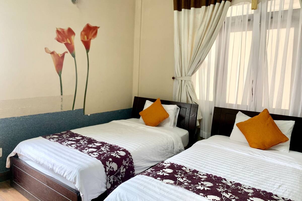 Une chambre d’hôtel avec deux lits, chacun agrémenté de draps blancs et d’oreillers orange, un mur décoré d’une fresque florale et une fenêtre avec des rideaux blancs.