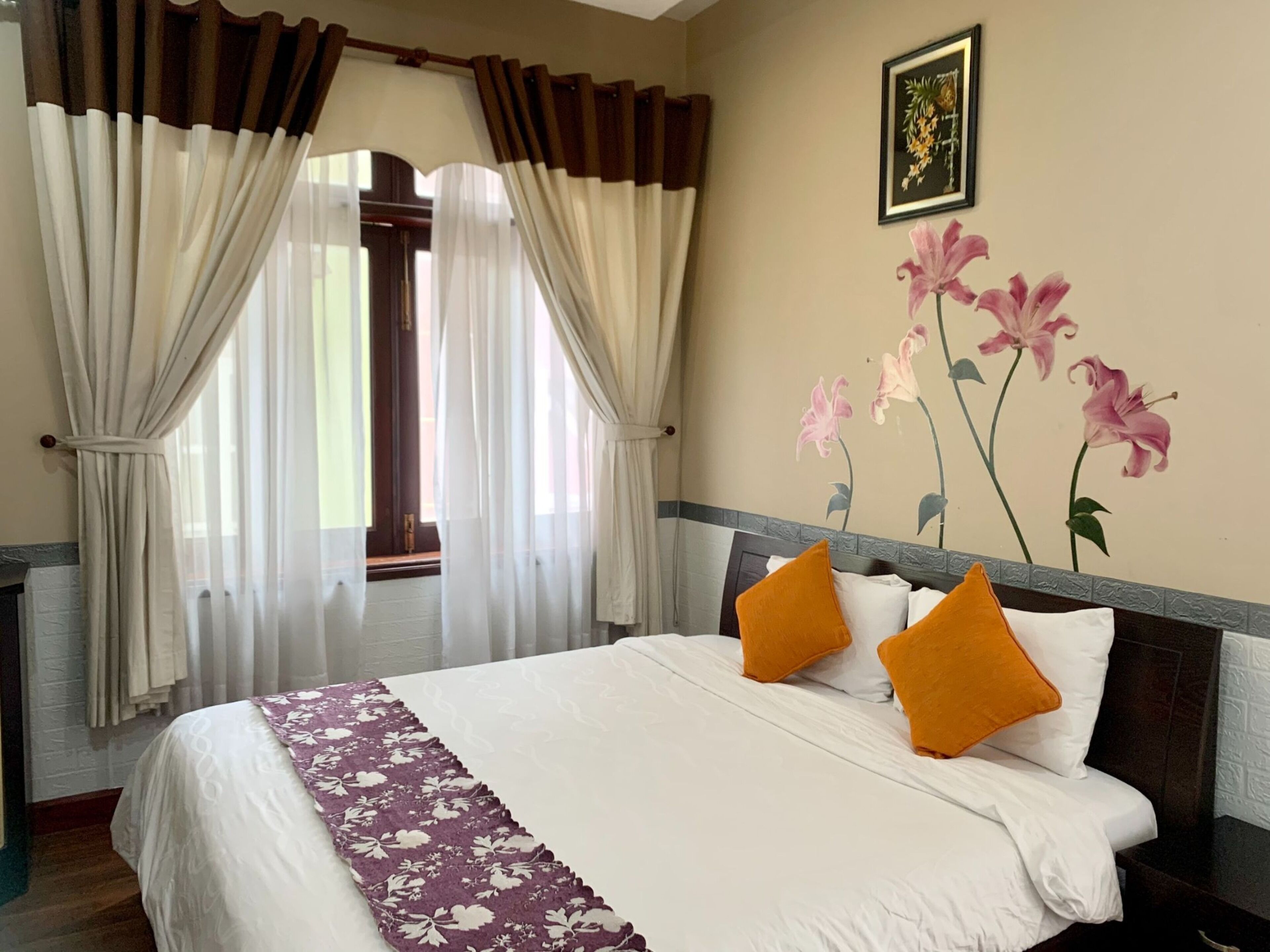 A hotel room with a bed, curtains, a floral wall decoration, and a framed picture.