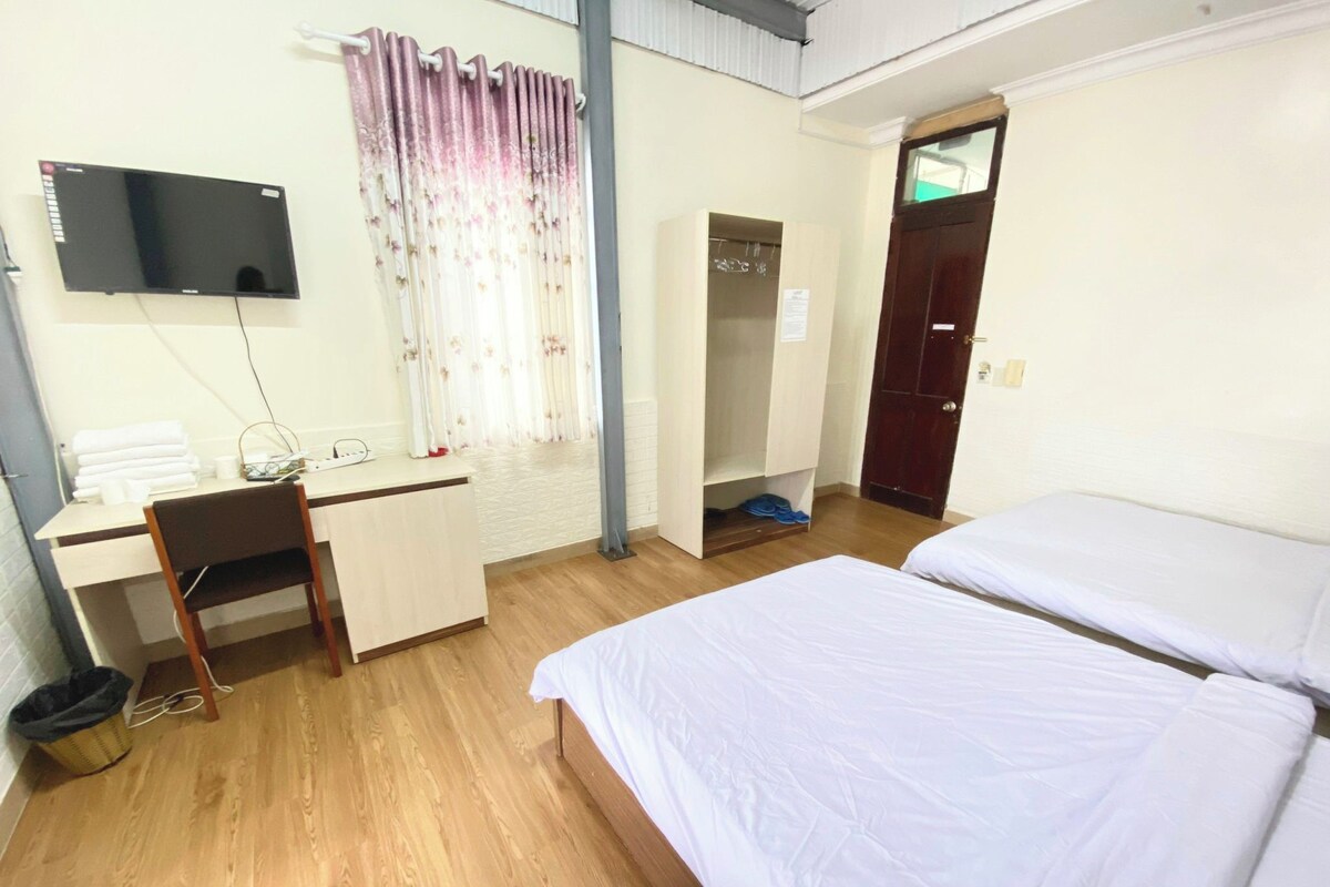 A hotel room with a bed, a desk with a chair, a television mounted on the wall, and a wardrobe.