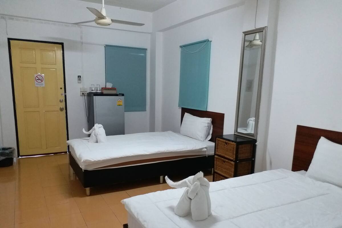 Quarto de hotel com duas camas de solteiro, uma mesa de cabeceira de madeira, um espelho, um ventilador de teto e uma porta com o aviso ‘proibido fumar'.