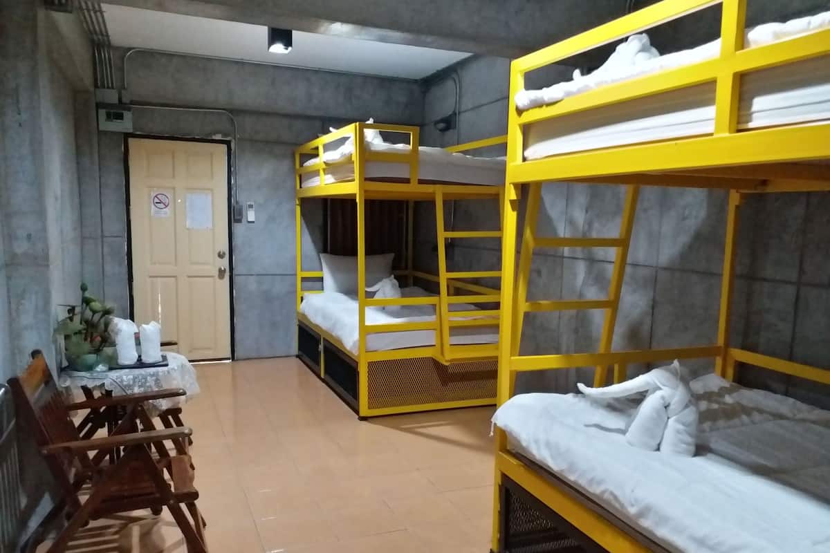 Um quarto de dormitório com beliches, uma cadeira de madeira, uma mesinha com toalha rendada e uma porta com placa de 'Não Fumar'.