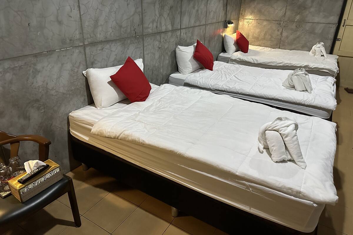 Quarto de hotel com três camas, cada uma com roupa de cama branca e almofadas vermelhas. Há uma mesa de cabeceira com um controle remoto e um estojo de lenços.