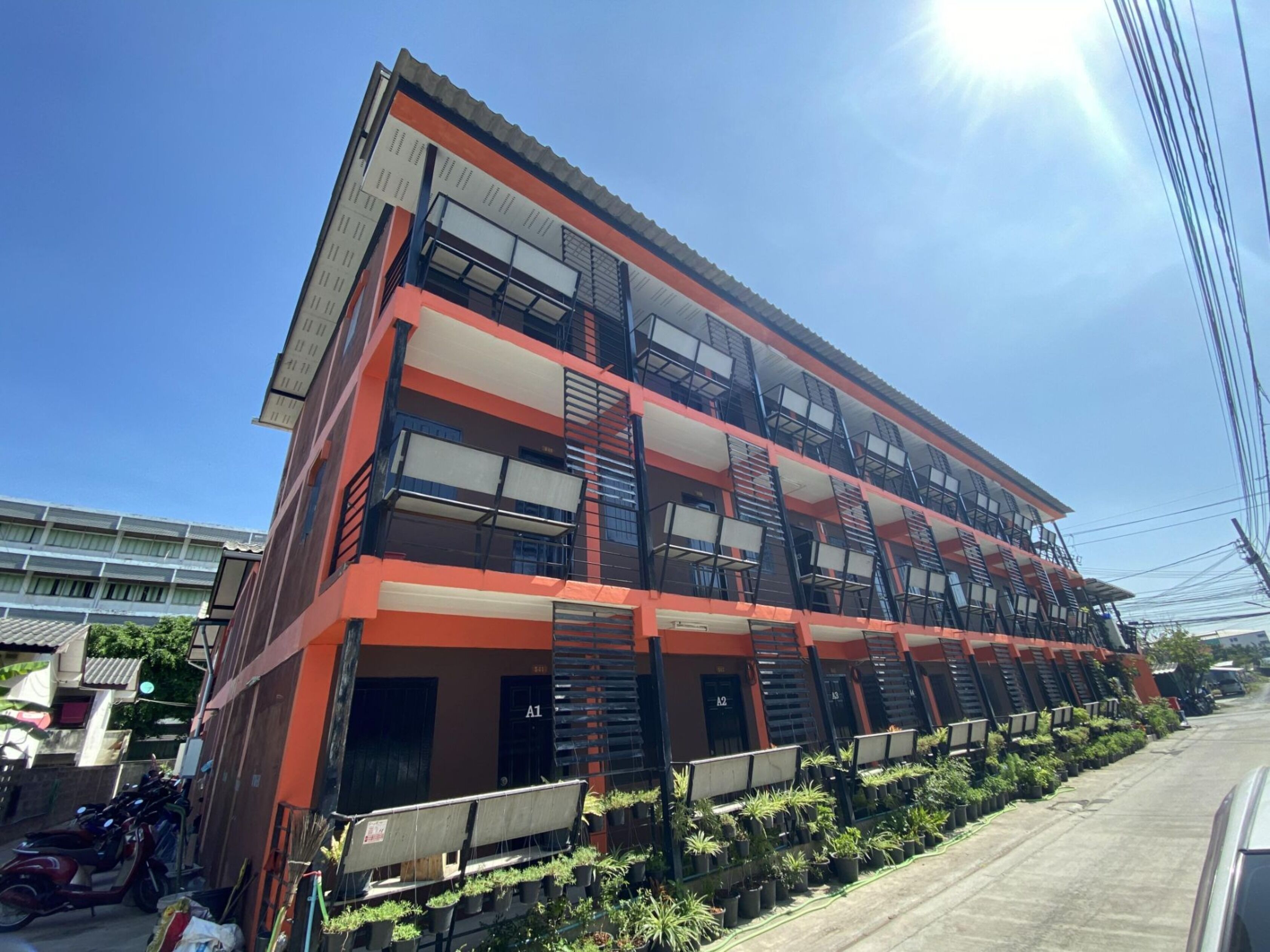 A multi-story building with balconies, a parking area with parked vehicles, and a row of potted plants along the sidewalk.