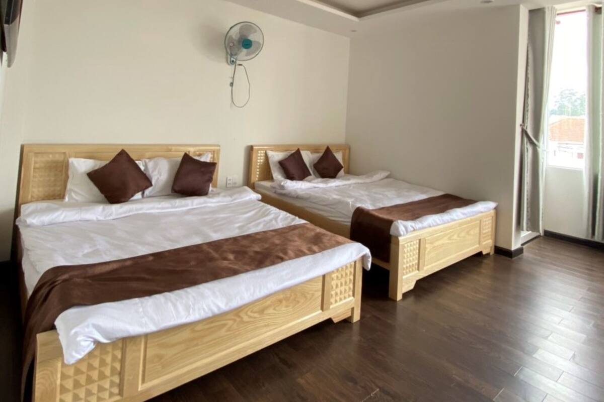 Two wooden beds with white and brown bedding, a wall-mounted fan, and a large window with a view of buildings outside.
