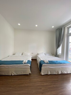 Habitación de hotel con dos camas individuales, suelo de madera, un ventanal grande con cortinas y pared blanca.