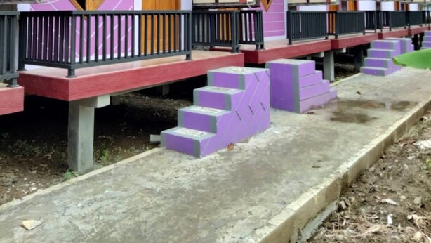 Een rij kleurrijke, rechthoekige chalets met paarse en roze gevels, houten deuren en een betonnen pad dat naar de ingang leidt.